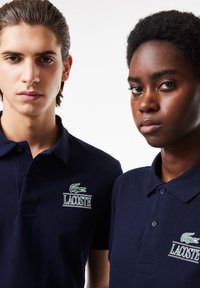 Dos personas llevan camisetas polo azul marino con el logo de Lacoste en verde. La tela parece texturizada con un diseño clásico y cuello.