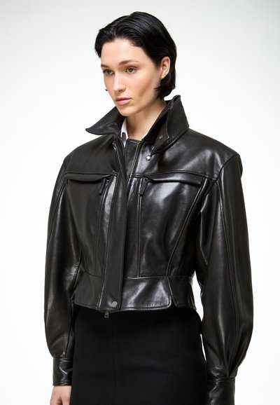 Žena v priliehavej čiernej koženej bunde s vysokým golierom, predným zipsom, patentnými gombíkmi a tvarovanými dlhými rukávmi, oblečená cez čiernu sukňu.