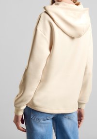 Kremowa bluza z kapturem o luźnym kroju, z regulowanym kapturem i ściągaczami na mankietach, zestawiona z jasnoniebieskimi dżinsami.