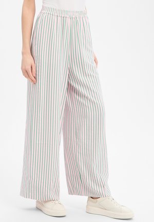 Femme portant un pantalon à jambes larges à rayures verticales vertes et des baskets blanches, debout devant un fond blanc uni.