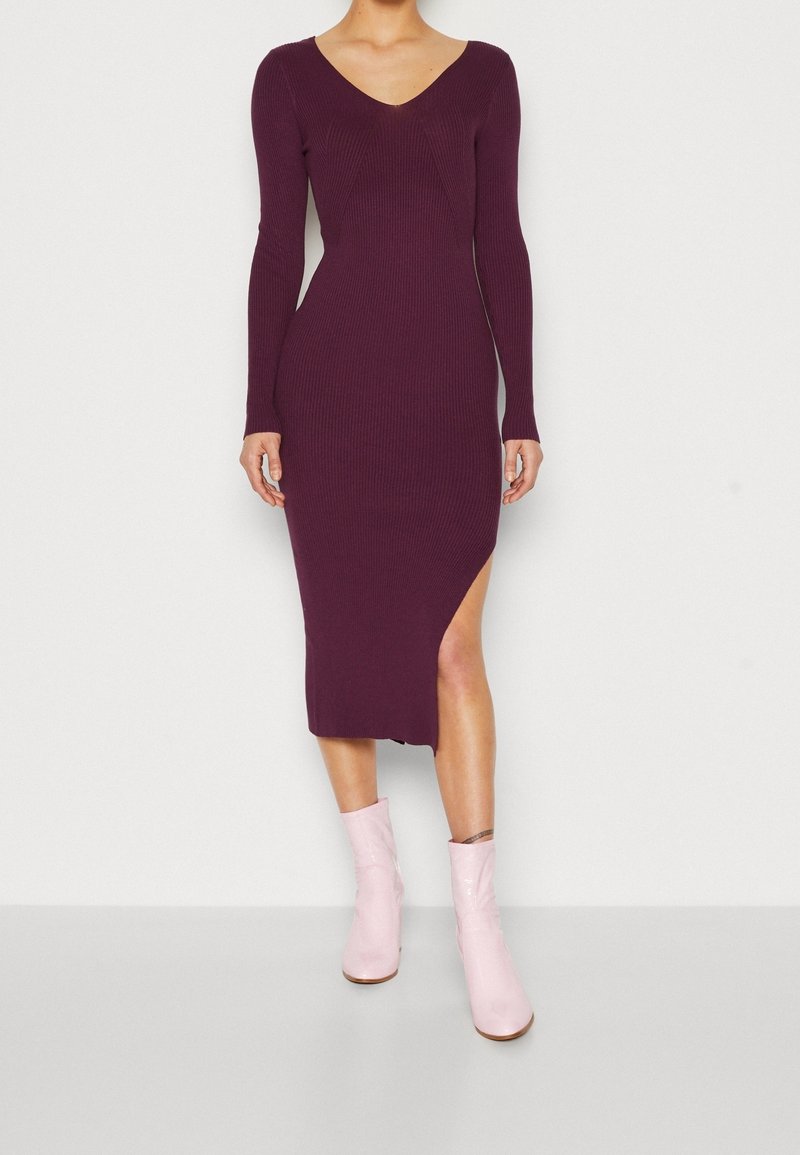 Femme portant une robe midi côtelée à manches longues, de couleur violet foncé, avec une fente latérale, associée à des bottines roses claires, debout devant un fond blanc.