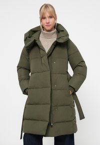 Puffer coat in olive green, featuring a hood, snap and zipper closure, and a quilted design. Soft fabric with side pockets.