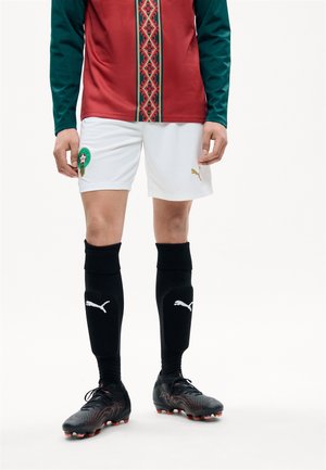 Ποδοσφαιριστής που φοράει λευκό σορτς με πράσινο-κόκκινο-χρυσό έμβλημα, μαύρες κάλτσες Puma, μαύρα παπούτσια και κόκκινη-πράσινη μακριά μπλούζα.