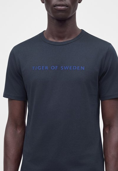 Άνδρας που φοράει μπλε ναυτικό t-shirt με κοντά μανίκια και την επιγραφή "TIGER OF SWEDEN" με μπλε γράμματα στο στήθος.