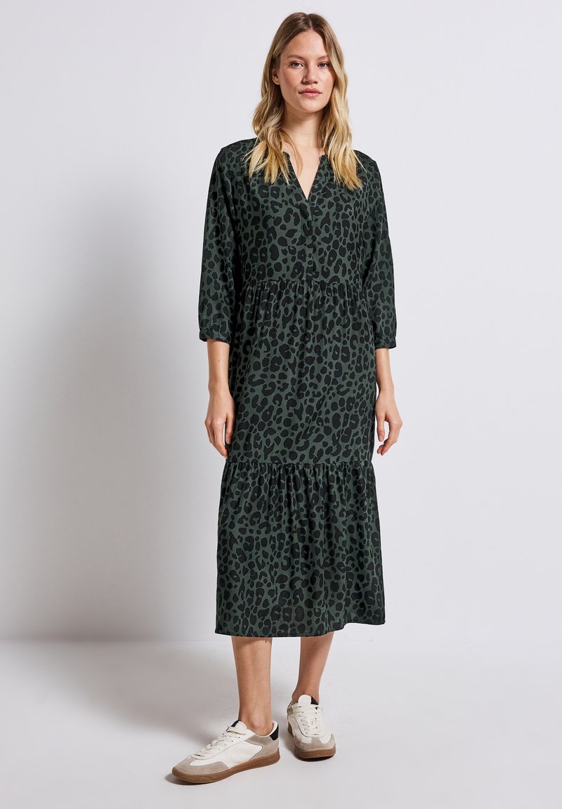 Grön leopardtryckt midiklänning med trekvartsärmar, lager av volanger vid fållen, i kombination med neutrala sneakers. Slät tygtextur.