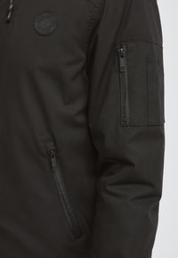 Schwarze Jacke aus glattem Stoff mit einer Reißverschlusstasche am Ärmel und einem runden Logo-Patch auf der Brust.