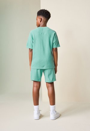 Dječak stoji okrenut leđima, nosi majicu kratkih rukava i kratke hlače u boji metvica, bijele visoke čarape i tenisice.