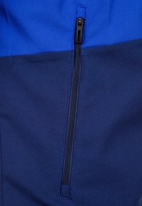 Blaue Jacke mit einem Reißverschlußfach. Der obere Teil ist hellblau, während der untere Teil dunkelblau und glatt ist.