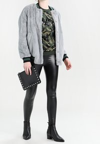 Giacca bomber in pelliccia sintetica grigia, polsini a coste verdi, felpa con grafica camouflage, leggings neri lucidi, pochette nera con borchie, stivaletti con punta.