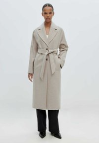 Lange beige wollen jas met een brede kraag, knoopsluiting en strikceintuur. Draagt over zwarte wijde broek en puntige zwarte schoenen.