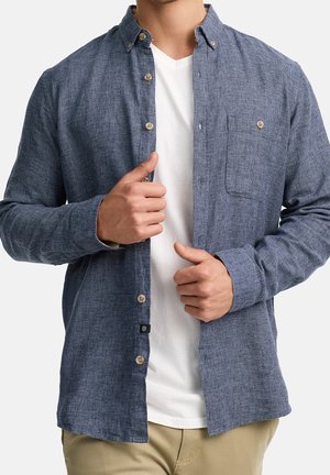 Mężczyzna w rozpiętej niebieskiej teksturowanej koszuli z długim rękawem na białym T-shircie z dekoltem w serek i beżowych spodniach, poprawia przód koszuli obiema rękami.