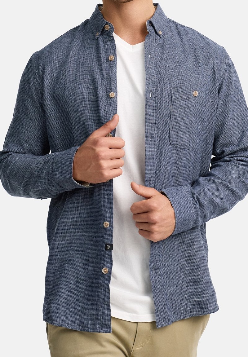Homme portant une chemise bleu texturée à manches longues déboutonnée par-dessus un t-shirt blanc à col en V et un pantalon beige, ajustant l'avant de la chemise avec ses deux mains.