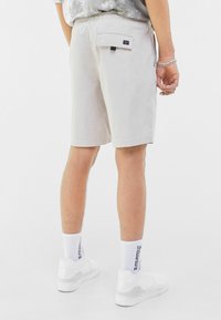 Shorts beige clair avec une taille élastique, une poche arrière, et une étiquette visible. Associés à des baskets blanches et des chaussettes avec du texte.