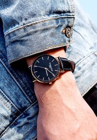 Reloj negro con detalles en dorado sobre una correa de cuero negro, llevado en una muñeca; manga de chaqueta de denim visible con detalle de costura.