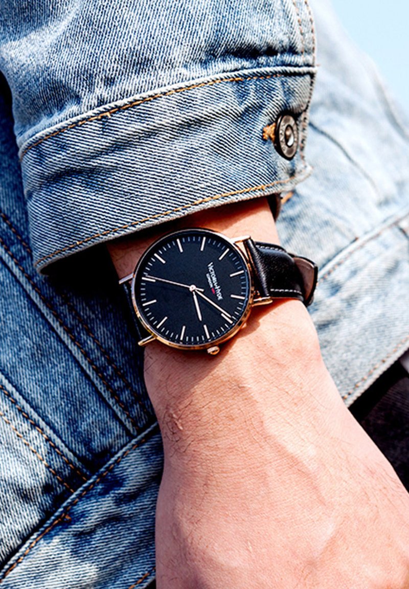 Reloj negro con detalles en dorado sobre una correa de cuero negro, llevado en una muñeca; manga de chaqueta de denim visible con detalle de costura.