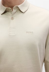 Μπεζ πόλο πουκάμισο κατασκευασμένο από απαλό ύφασμα, με γιακά, τρεις κουμπότρυπες και διακριτικό κεντητό λογότυπο της μάρκας στο στήθος.