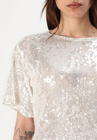 Top bianco trasparente con tessuto testurizzato e paillette, scollatura ampia e maniche corte. Strisce sottili visibili nel materiale.