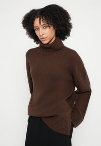 Pulover brun cu guler înalt, tricotat cu textură nervurată, cu deschideri laterale și croială lejeră. Mâneci lungi și tiv ușor curbat.