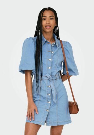 Denim obleka v svetlo modri barvi, z napihnjenimi rokavi, sprednjim delom z gumbi, pasom in dolžino do kolen. Majhna rjava torbica čez ramo.