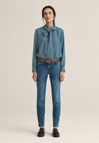 Lyseblå bluse med sløjfe, parret med tætsiddende mørkeblå jeans og sorte loafers. Har glat stof og et klassisk design.