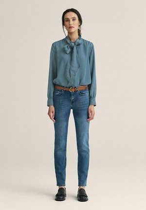 Lyseblå bluse med sløjfe, parret med tætsiddende mørkeblå jeans og sorte loafers. Har glat stof og et klassisk design.