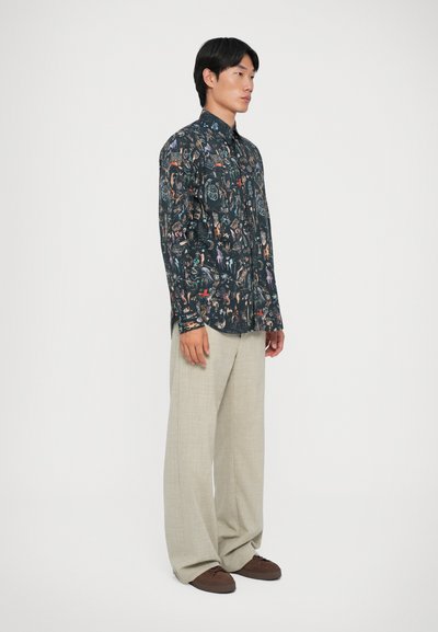 Camicia a maniche lunghe con bottoni in tessuto scuro, caratterizzata da motivi multicolore. Abbinata a pantaloni a gamba larga beige chiaro e scarpe marroni.