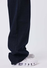 Pantalones de denim de pierna ancha en azul marino, con una costura única en el lateral y combinados con zapatillas deportivas blancas y gruesas.