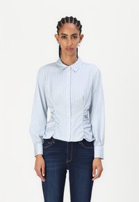 Lyseblå stripete bluse med knapper foran, krage og samlet sidedele. Lange ermer med knapper i mansjettene. Brukt sammen med mørke jeans.