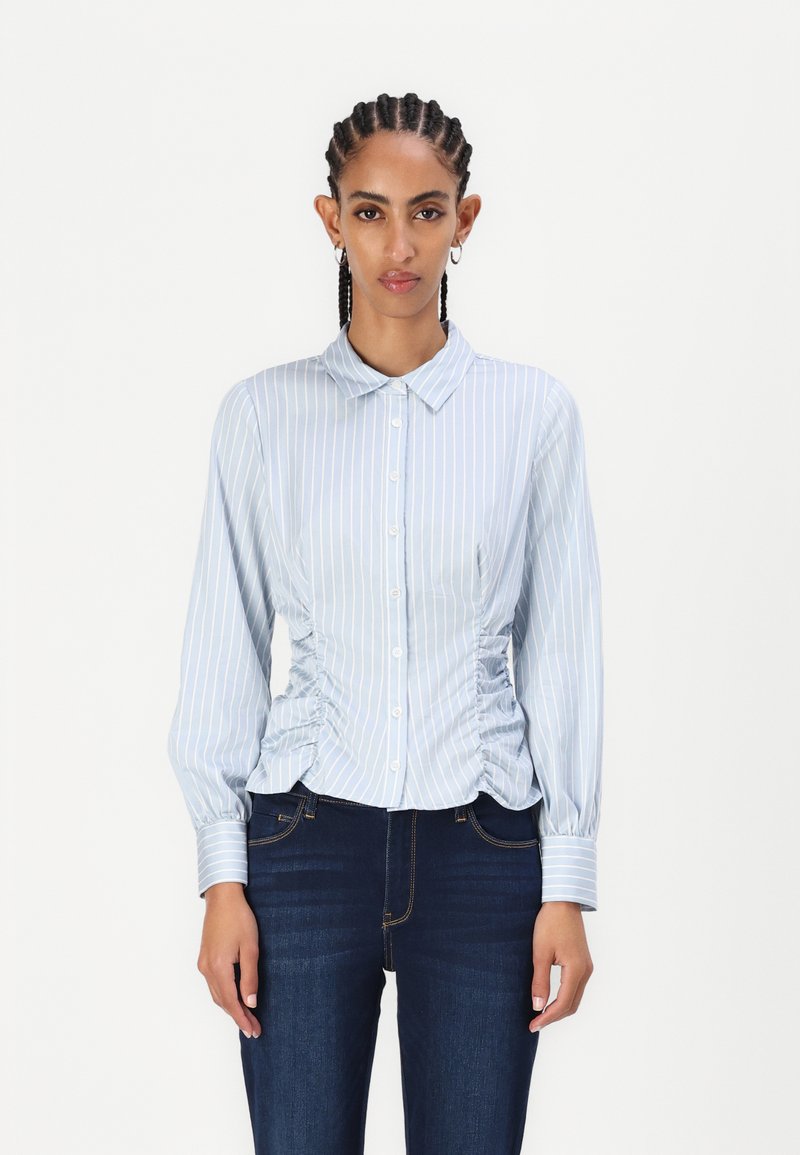 Lyseblå stripete bluse med knapper foran, krage og samlet sidedele. Lange ermer med knapper i mansjettene. Brukt sammen med mørke jeans.