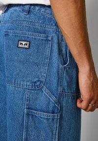 Blue denim cargo pants featuring multiple pockets, contrasting stitching, and a rectangular black logo patch on the back pocket.