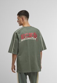 Olijfgroen oversized t-shirt met een grote rode en witte graphic op de achterkant en contrasterende stiksels langs de mouwen.