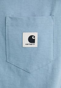 Poche en tissu bleu clair avec un patch en carré blanc de logo Carhartt cousu au centre de la poche.