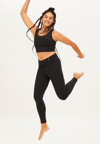 Zwarte sportieve crop top en legging, met een gestructureerd ontwerp en een getailleerde pasvorm. Het model springt blootsvoets tegen een lichte achtergrond.