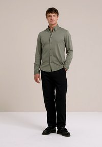 Junger Mann, der mit teilweise in die Taschen gesteckten Händen steht, trägt ein grünes, geknöpftes Shirt, schwarze Hosen und schwarze Schuhe vor einem einfarbigen Hintergrund.