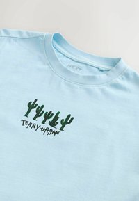 Tricou din bumbac de culoare albastră deschisă, cu ilustrații verzi de cactuși și textul "TERRY URBAN". Design cu guler rotund, mâneci scurte și croială obișnuită.