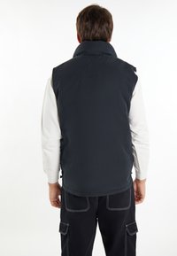 Gilet imbottito blu navy con colletto alto, texture liscia e dettagli di cucitura orizzontale, indossato sopra una camicia bianca a maniche lunghe.