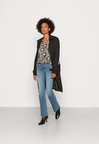 Um casaco trench preto sobre uma blusa floral em preto e branco, combinado com jeans flare azul claro e botas pretas de bico fino.