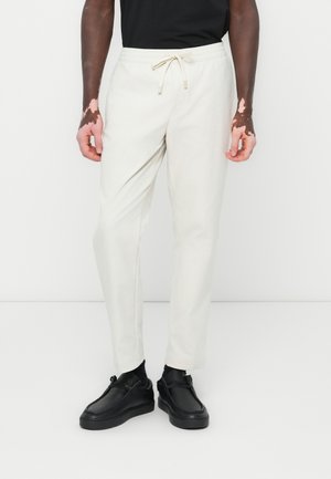 White tapered jogger pants with an elastic waist and drawstring. Smooth texture, featuring front pockets. Worn with black shoes.