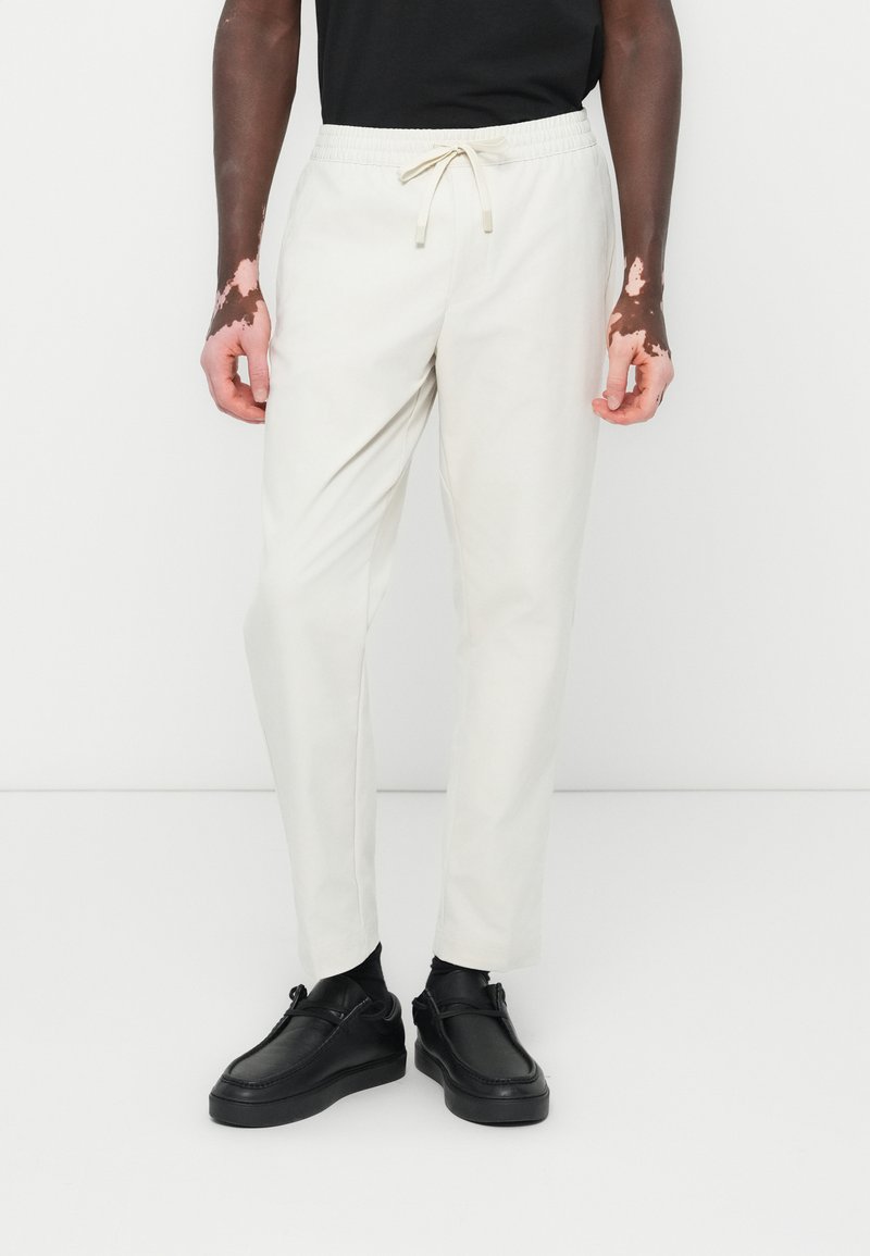 White tapered jogger pants with an elastic waist and drawstring. Smooth texture, featuring front pockets. Worn with black shoes.