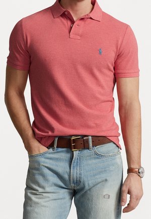 Homme portant un polo rouge à manches courtes avec un petit logo, un jean bleu clair, une ceinture en cuir marron et une montre-bracelet.
