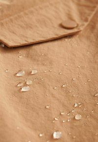 Gouttes d'eau perlées sur un tissu imperméable beige avec un rabat boutonné en arrière-plan.