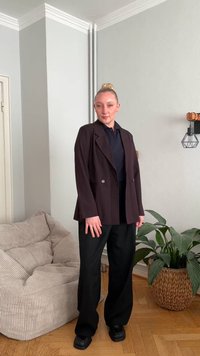 Tumšs pinstripe žakete pāri tumši zilam polera krekliņam, papildināta ar plašām melnām biksēm un masīvām melnām kurpēm. Gaiša tekstūra un neitrālas fona detaļas.