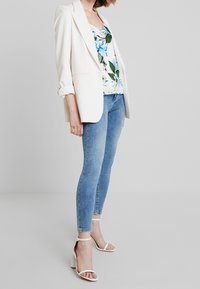 Vit blazer över blommig linne, kombinerad med blå snäva jeans och vita klacksandaler. Slät tyg; avslappnad passform; uppvikta blazerärmar.