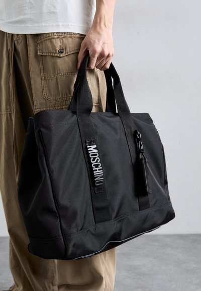 Mustmust kott mustast kangast, kahekordsete lühikeste käepidemetega, lukuga sulgemine, silmatorkav "Moschino" logo ja tekstuuriga pind.