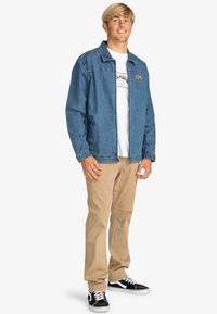 Denim jacket in light blue, featuring a collar and front pockets. Worn over a white t-shirt and paired with beige pants and black sneakers.