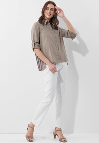 Khakifarbenes Hemd mit aufgerollten Ärmeln, gefalteter Rückseite, kombiniert mit weißen Hosen und beigen Absatzsandalen.