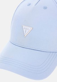 Gaiši zils beisbola cepures izgatavots no auduma, priekšpusē ar trīsstūra formas zelta logo, redzamām šuvēm un acīm augšpusē.