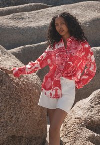 Camicia rossa a motivo con design astratti a tema marino; tessuto leggero. Abbinata a pantaloncini bianchi, posata contro rocce strutturate.