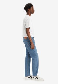 Camiseta blanca de manga corta combinada con jeans azules holgados que tienen un parche de cuero y zapatillas en blanco y negro. Composición de vista lateral.