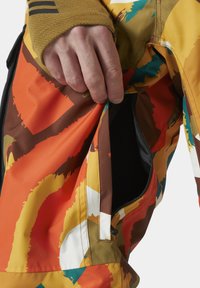 Barevné vzorované kalhoty v oranžových, hnědých a žlutých tónech, s kapsou na zip a texturovanou látkou pro zvýšení trvanlivosti.
